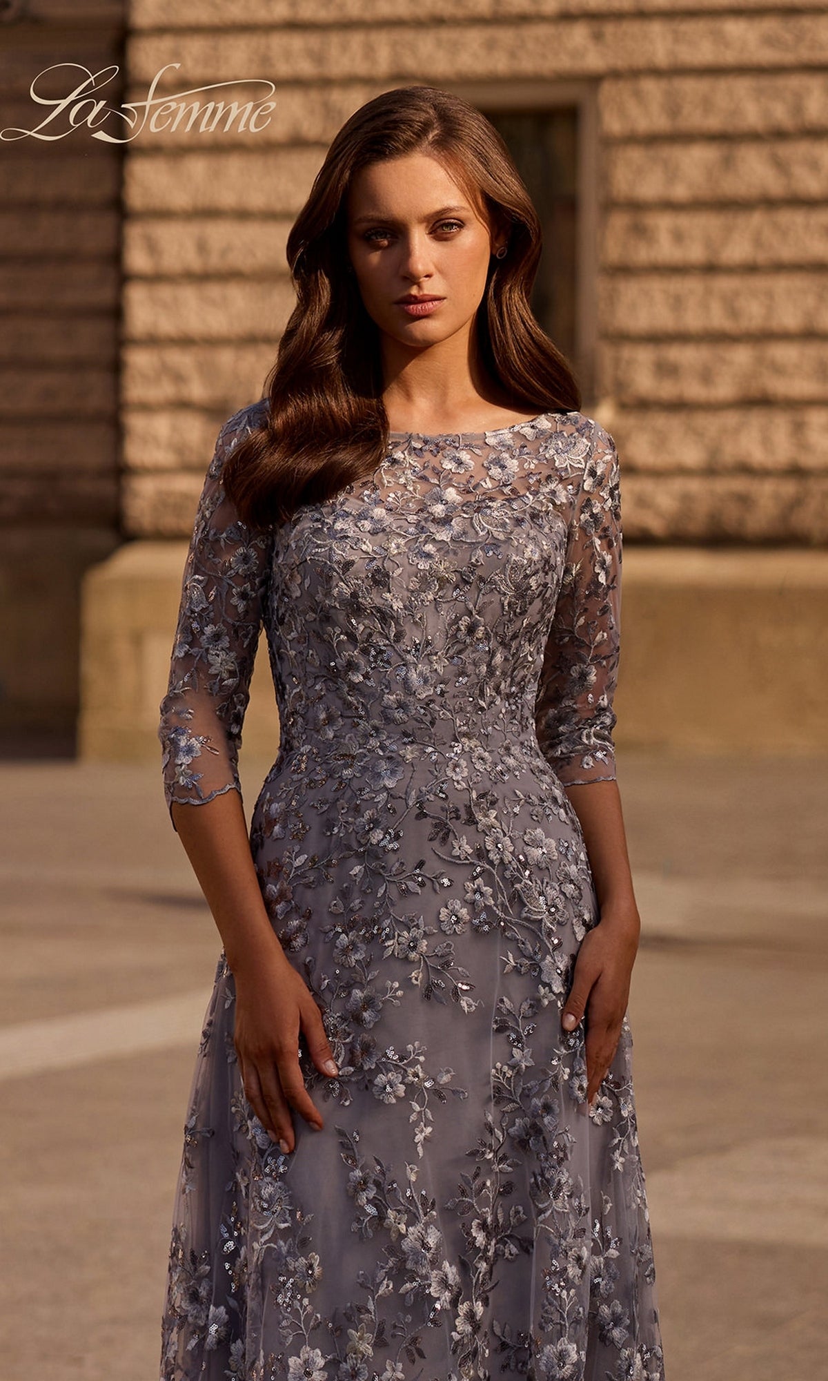 A woman in the Long Formal Dress: La Femme 29903, a silver gown with metallic floral lace and three-quarter sleeves, stands outside by a stone building.